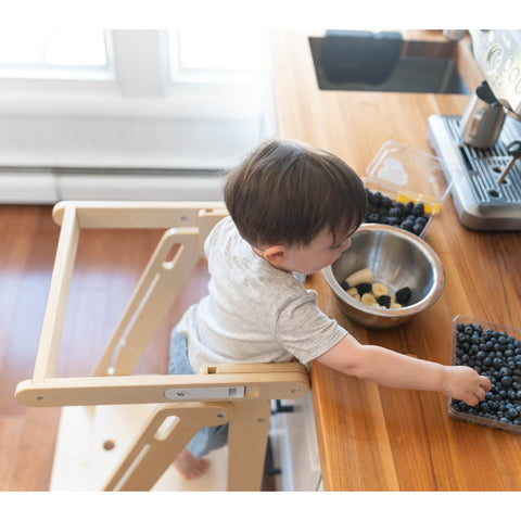 A young boy stands at the counter in the Learn N Fold Learning Tower in Natural color.