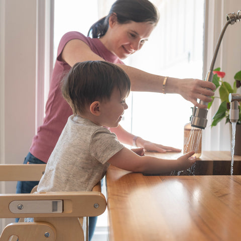 A young boy in the Learn N Fold Learning Tower and his mother are standing at the kitchen sink using the faucet.