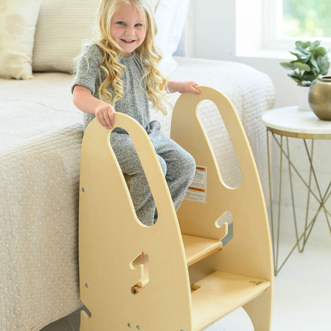 A young girl stands on the 3-in-1 Growing Step Stool near a bed.